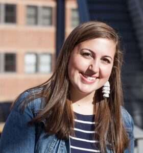 Picture of Lily, a young white woman with long hair in a jeans jacket, red brick building in the background