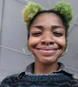 Picture of a person of color with green tufts of hair above black roots, silver nose ring, and big smile