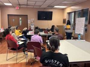 Picture of a group of 10 people in a classroom. A Black woman stands at a white board pointing to text on it.
