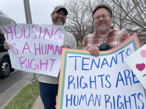 Picture of two men holding signs about housing and human rights outside on a sidewalk