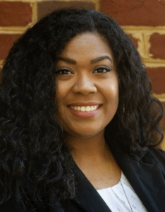Portrait photo of a woman of color with long curly black hair, white shirt, and black jacket